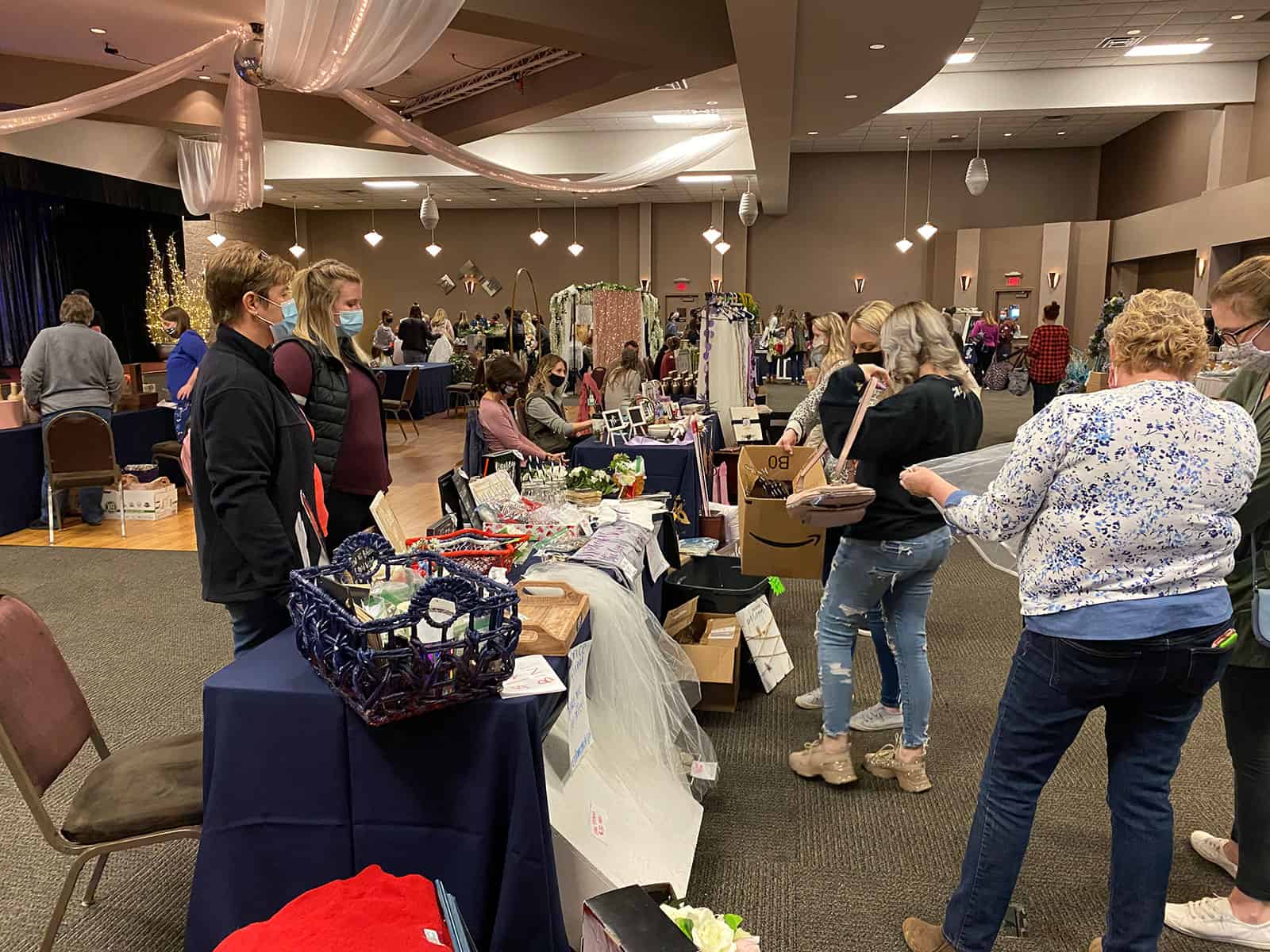 People browse and interact at wedding vendor tables in a large indoor event space decorated with draped fabric and lights.