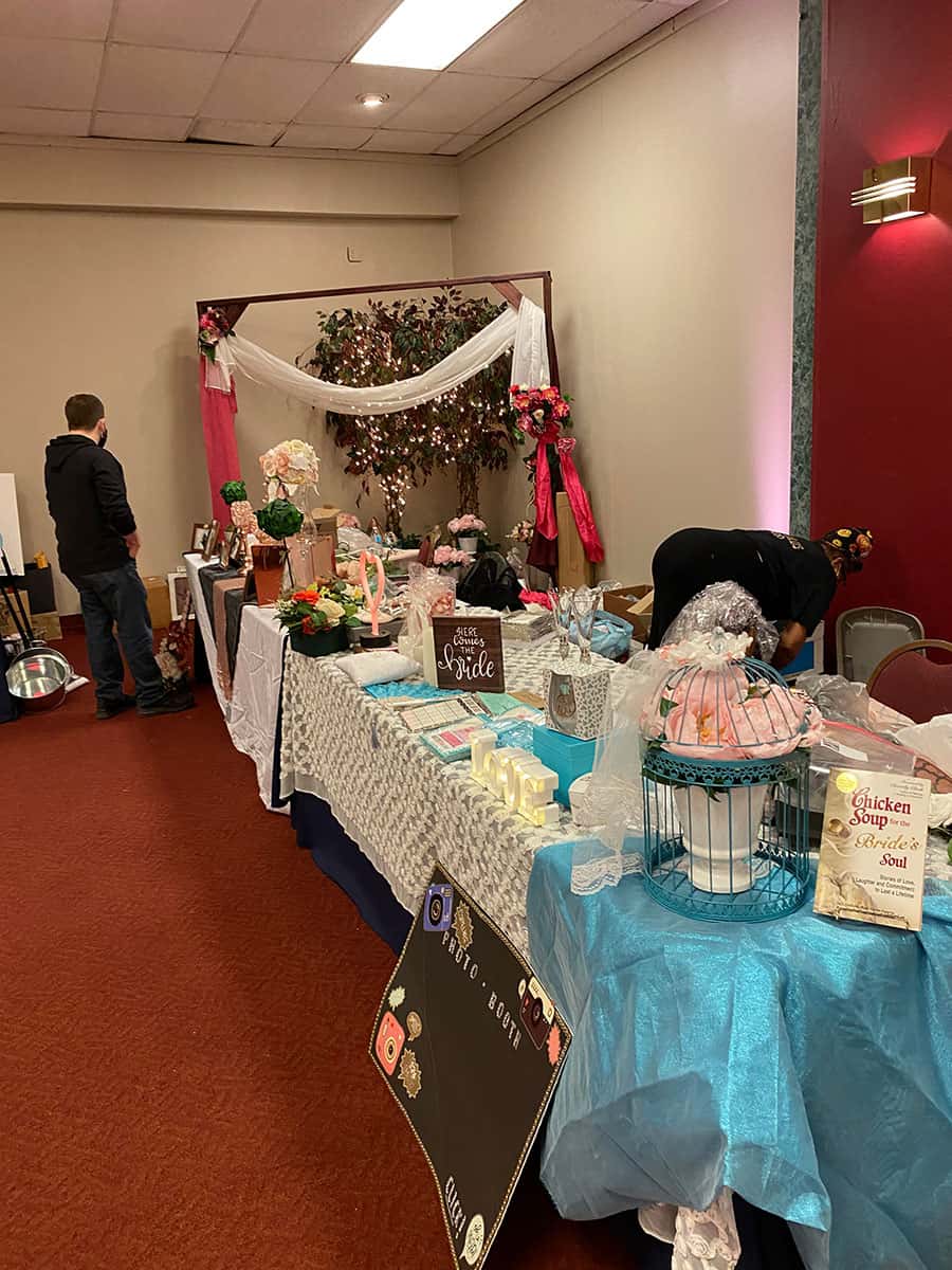 A decorated booth at an indoor bridal fair displays wedding-themed items, flowers, and gift baskets on tables; two people are present, one arranging items.