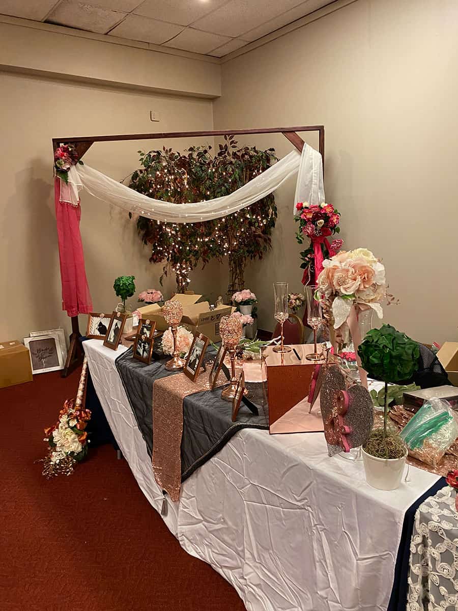 A decorated table with framed photos, flowers, and gift bags sits in front of an arch draped with white and pink fabric, with a lit plant in the background.