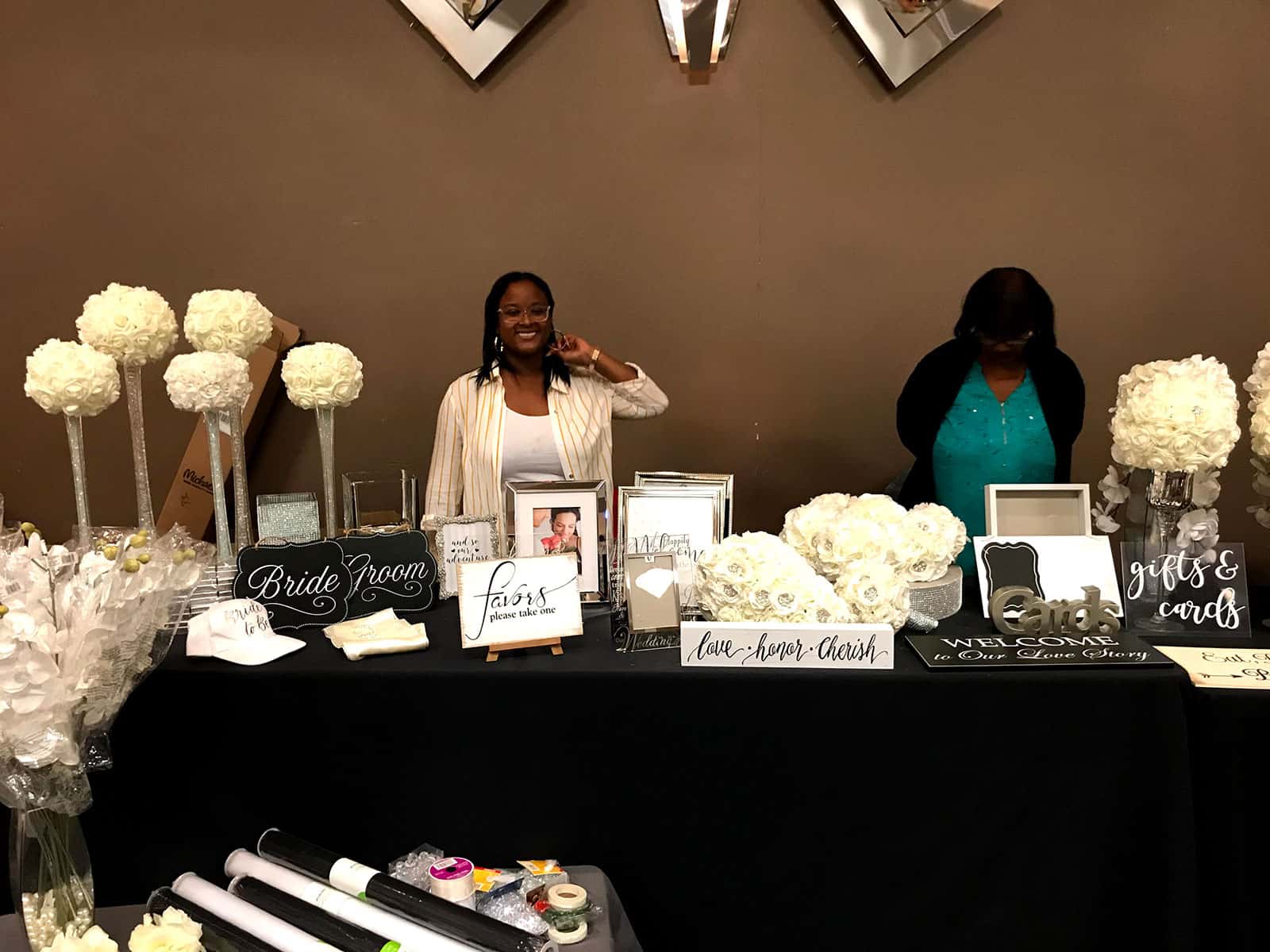Two women stand behind a booth displaying wedding décor items such as flowers, signs, and framed photos at a wedding show.