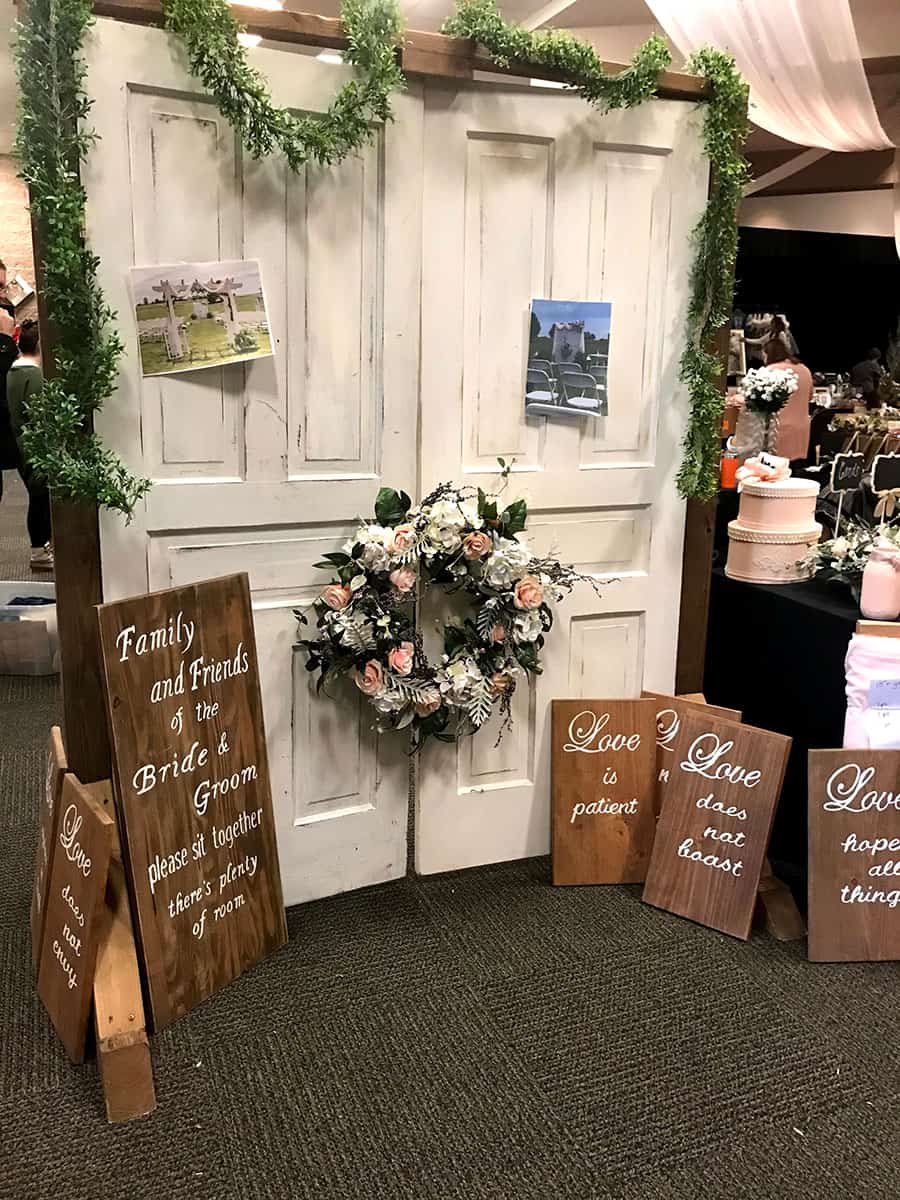 A decorative wedding display with white doors, floral wreath, greenery, and wooden signs with love quotes, set indoors on carpeted flooring.