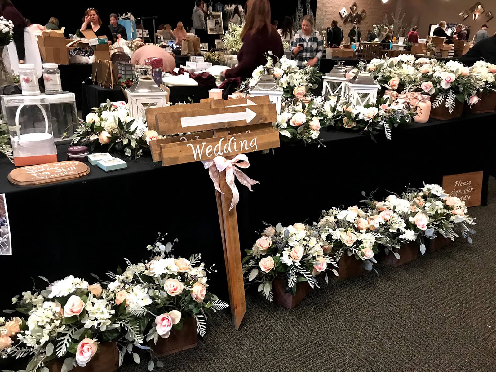 Decorated vendor table with floral arrangements, lanterns, and a "Wedding" sign with arrows pointing right. People browse in the background at an indoor bridal show.