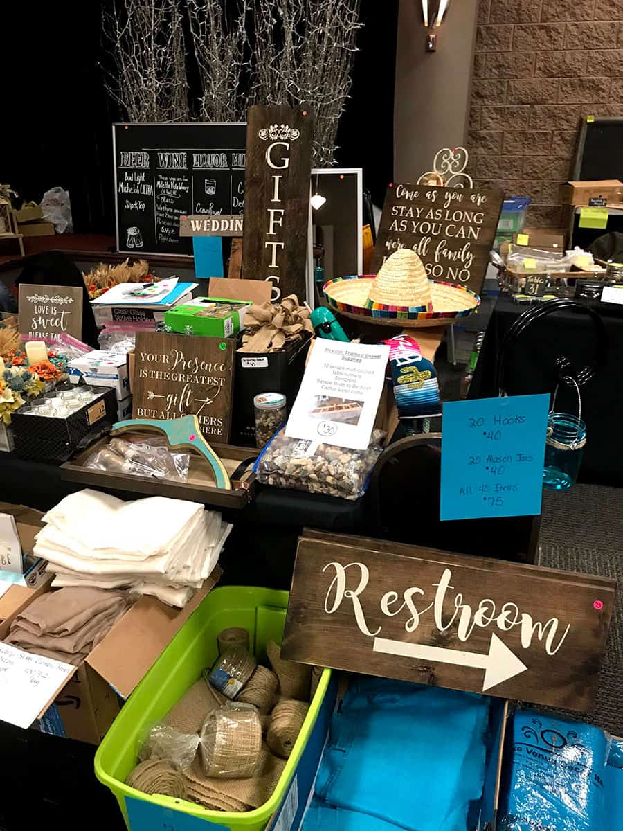A table displays assorted wedding decorations, signs, baskets, jars, and supplies for sale in a thrift or resale shop.