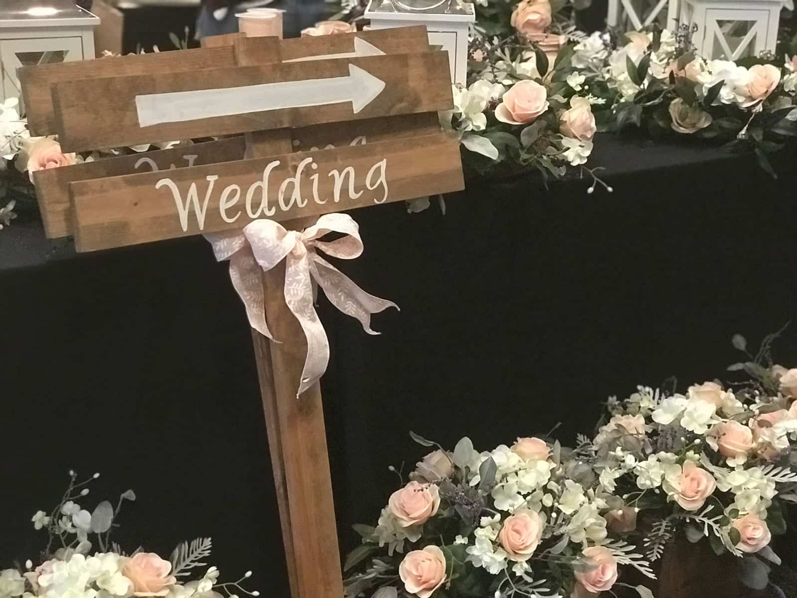 Wooden sign with an arrow and the word "Wedding" in white, surrounded by floral arrangements with pale pink and white roses.