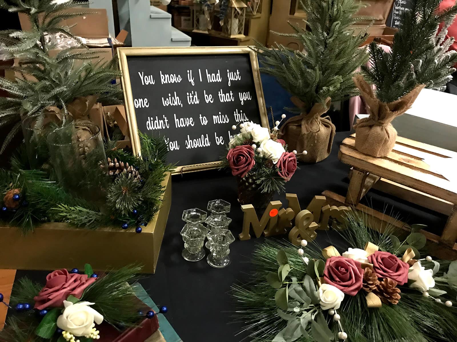Table displays faux plants, floral arrangements, candles, framed quote sign, small wooden decorations, and miniature Christmas trees.