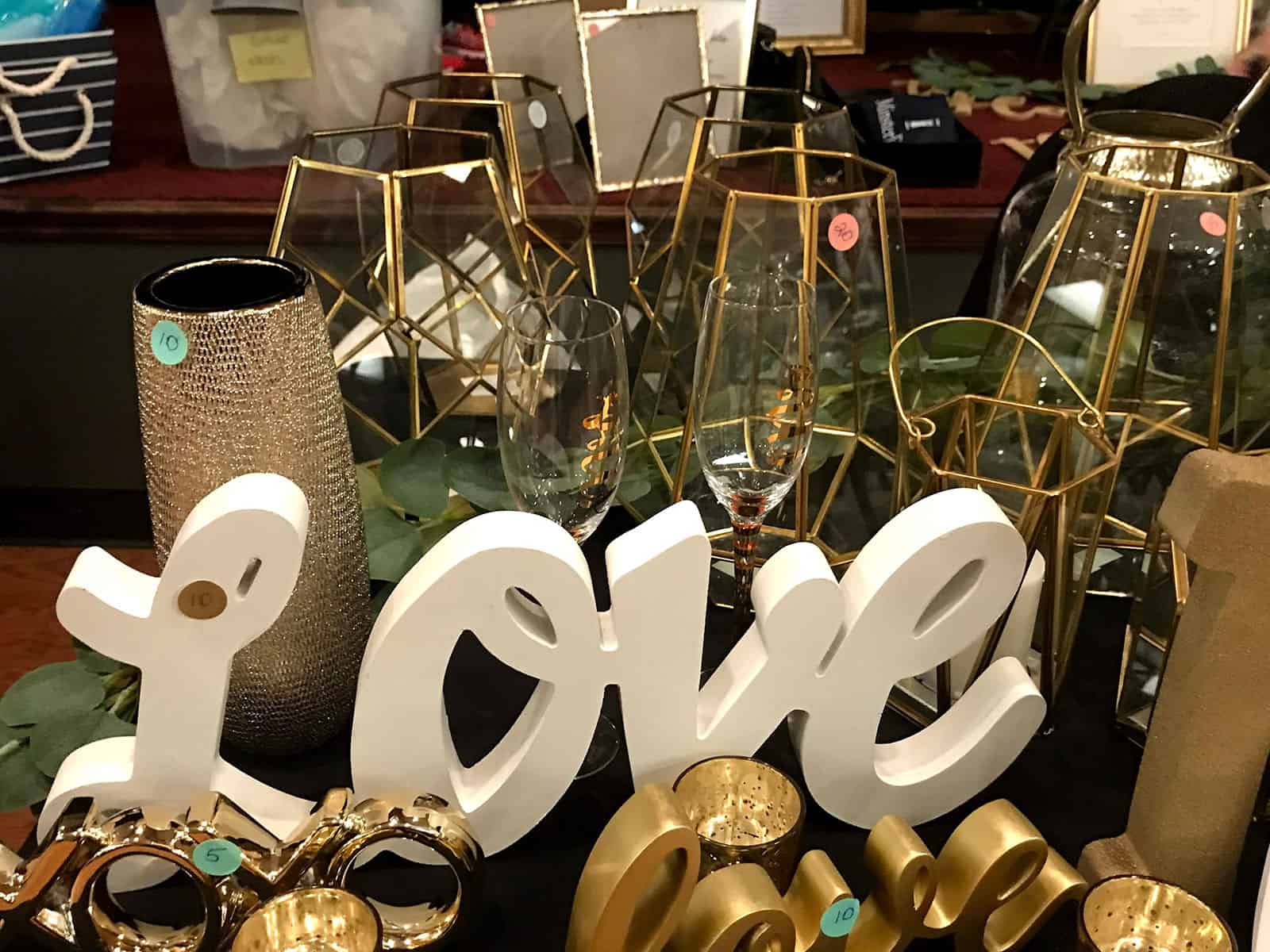 A tabletop display featuring gold candle holders, glass lanterns with gold frames, two champagne flutes, a glittery vase, and a large white "LOVE" sign.