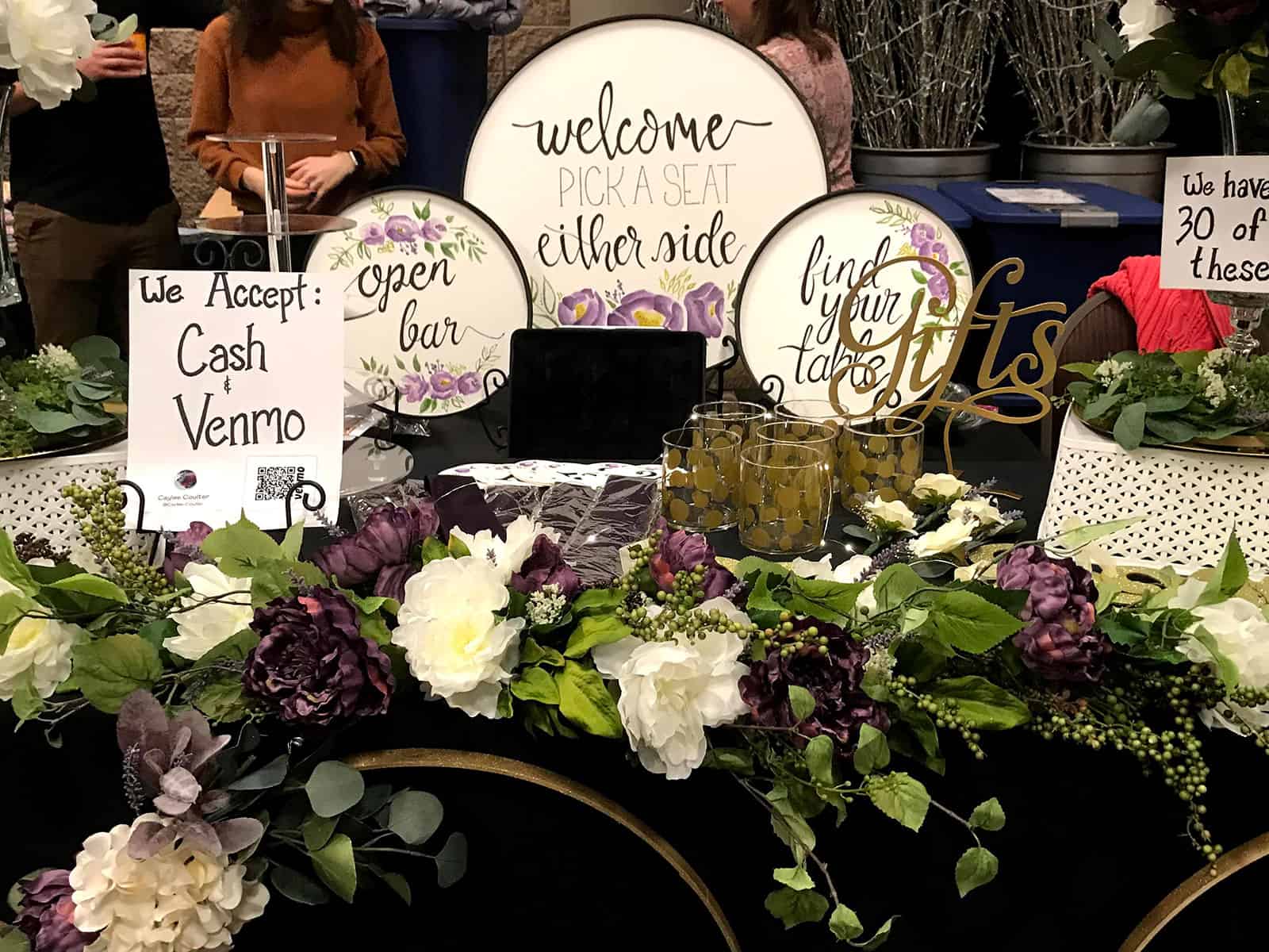 Decorated wedding table with floral arrangements, signs for seating and gifts, an “open bar” sign, and a note stating “We accept Cash and Venmo.”.