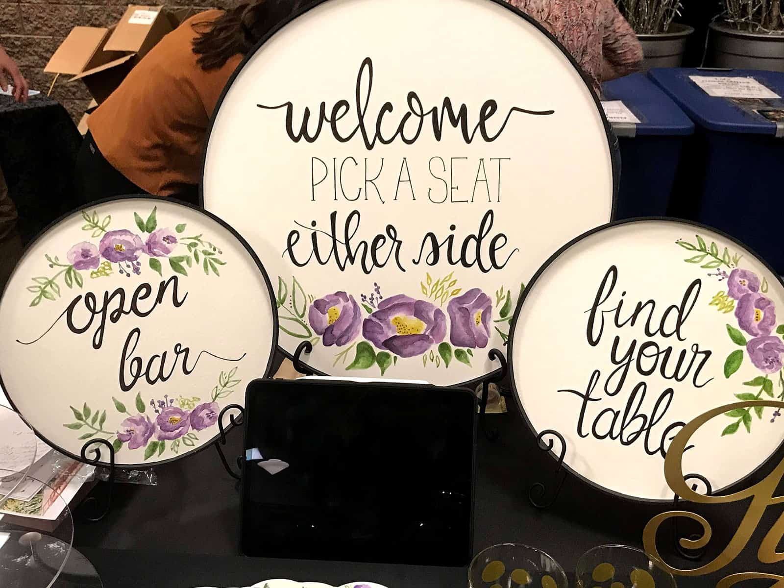 Three round signs with floral designs display the texts: "welcome pick a seat either side," "open bar," and "find your table".