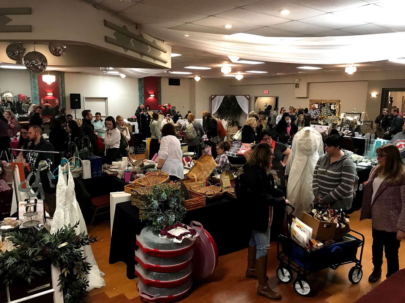 People browse various vendor booths at an indoor bridal expo event, with wedding dresses, baskets, and decorations on display.