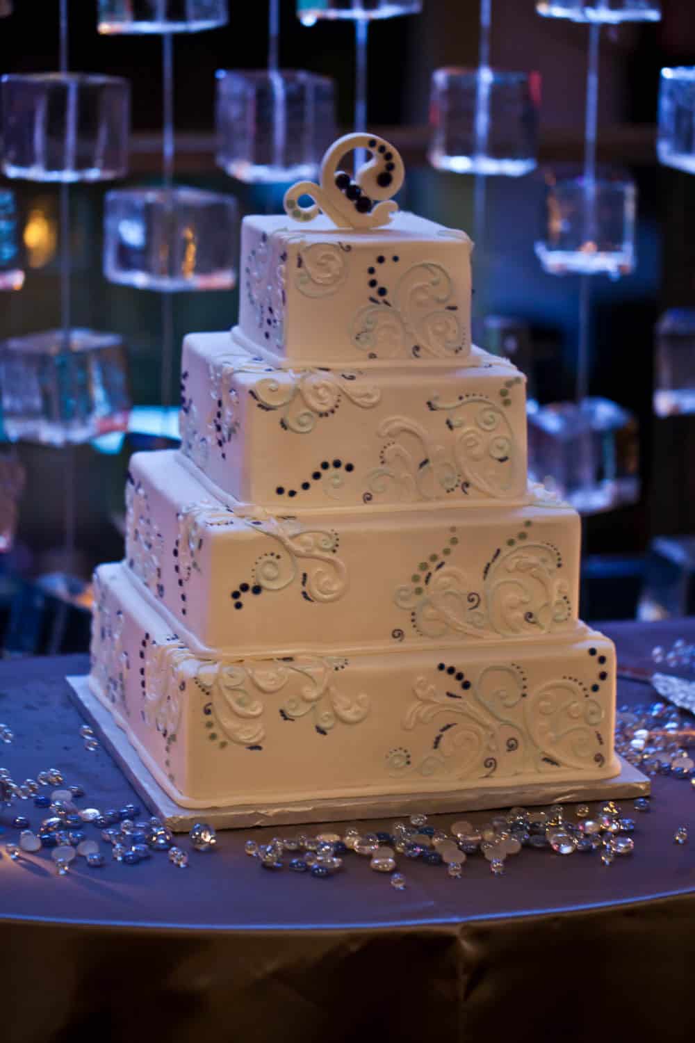 A four-tiered square wedding cake with white icing, blue and silver decorative swirls, and a circular topper, displayed on a table with scattered crystals.
