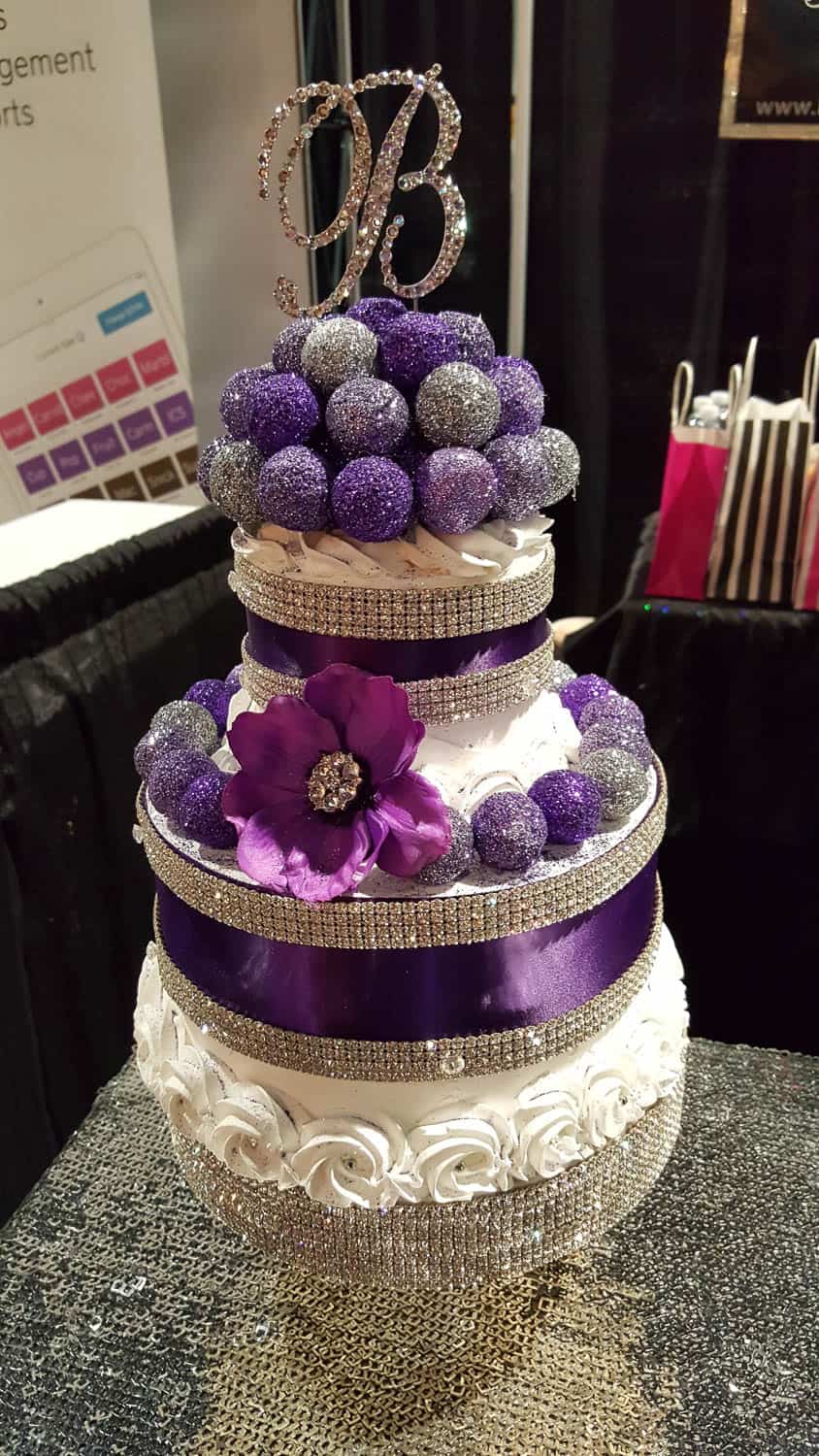 A three-tiered white cake decorated with purple and silver glittery spheres, a purple ribbon, a large purple flower, and a rhinestone "B" topper.