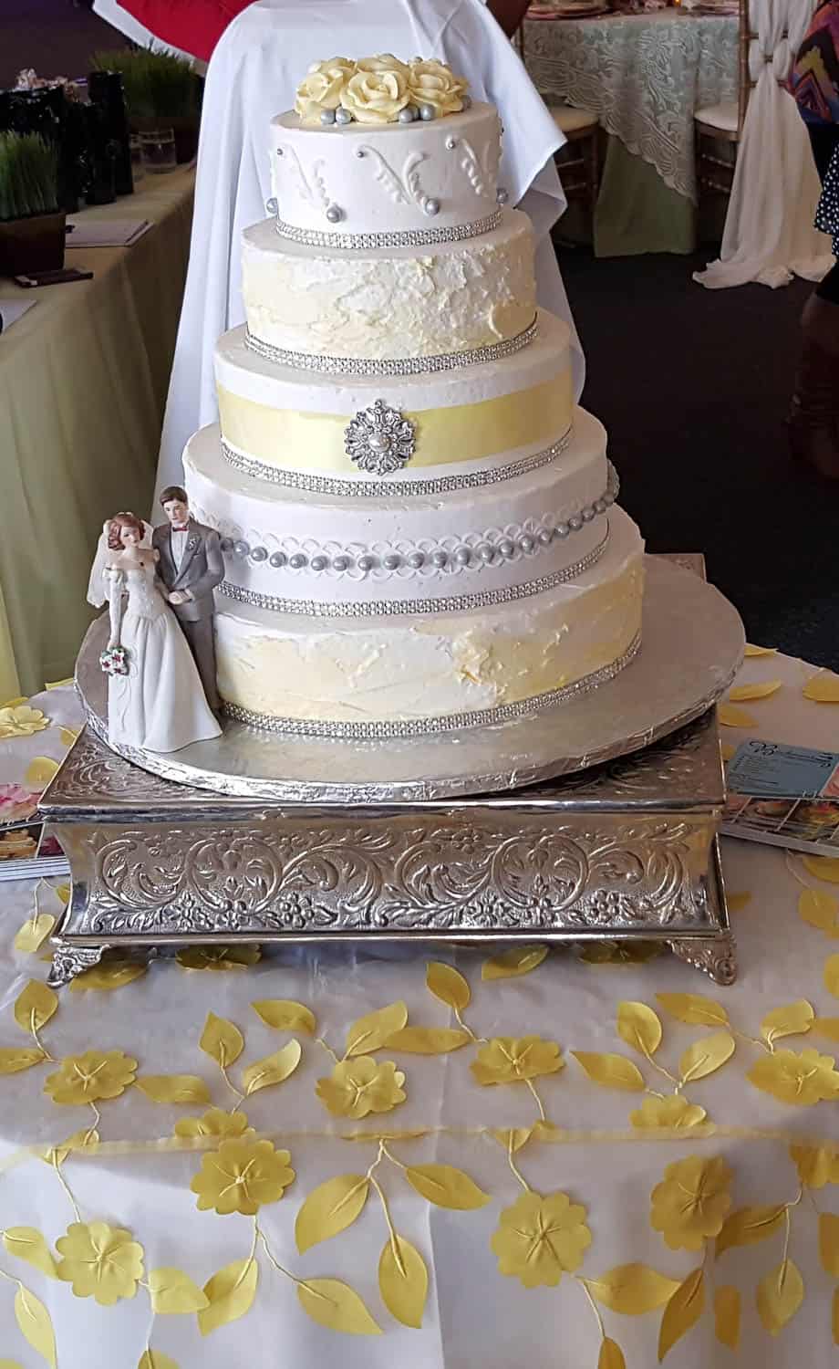 A five-tiered white and yellow wedding cake with a bride and groom topper, displayed on an ornate silver stand and decorated tablecloth.