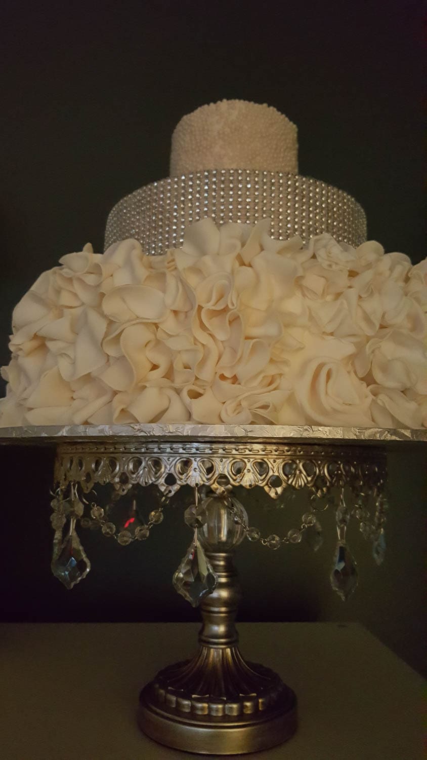 A three-tiered white cake with a ruffled lower tier and a beaded middle, on an ornate silver cake stand with crystal decorations.