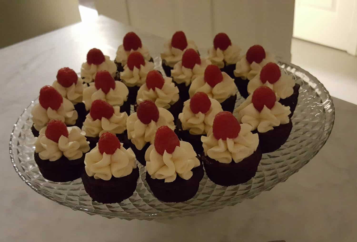 A glass platter with rows of small chocolate cupcakes, each topped with a swirl of white frosting and a single raspberry, sits on a marble surface.