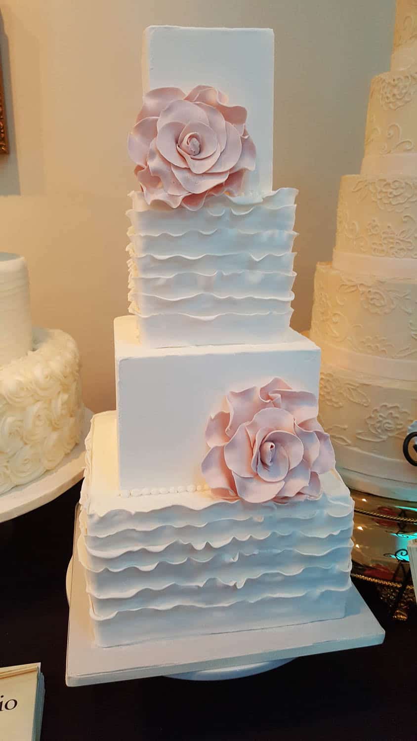 A four-tiered square white cake with ruffled icing and large pale pink fondant flowers on two tiers, displayed on a table.