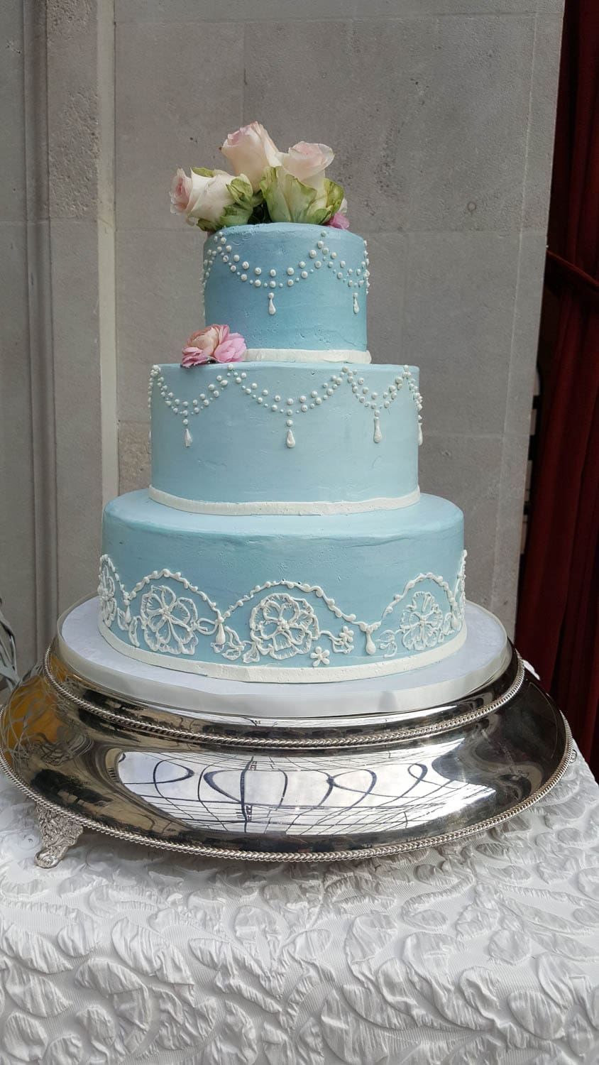 Three-tiered light blue cake with white icing decorations and pink roses on top, displayed on a silver tray against a light stone background.