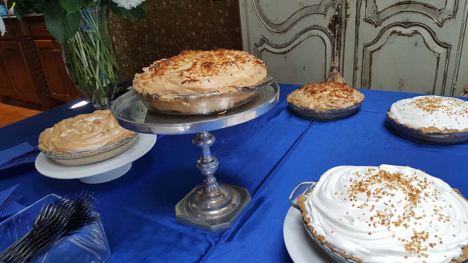 Five pies with different toppings are displayed on stands and plates on a blue tablecloth, with a vase of flowers and vintage doors in the background.