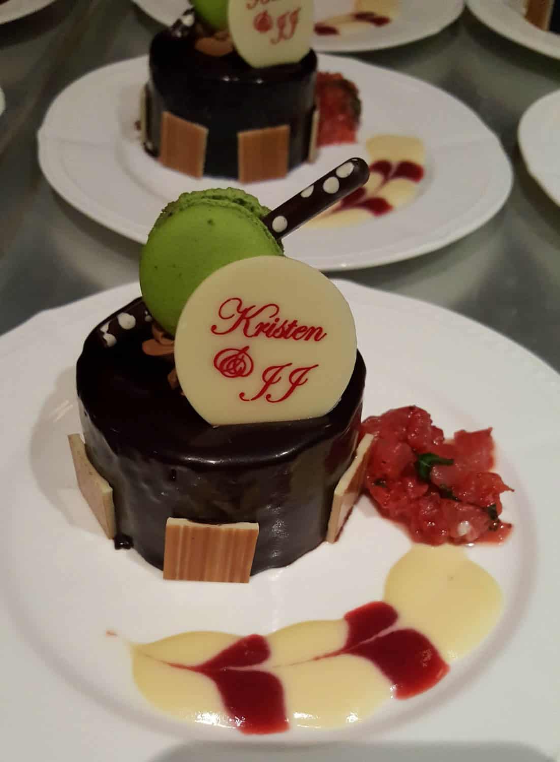 A chocolate dessert topped with a green macaron, a white chocolate disc reading "Kristen and JJ," and decorative sauces on a white plate.