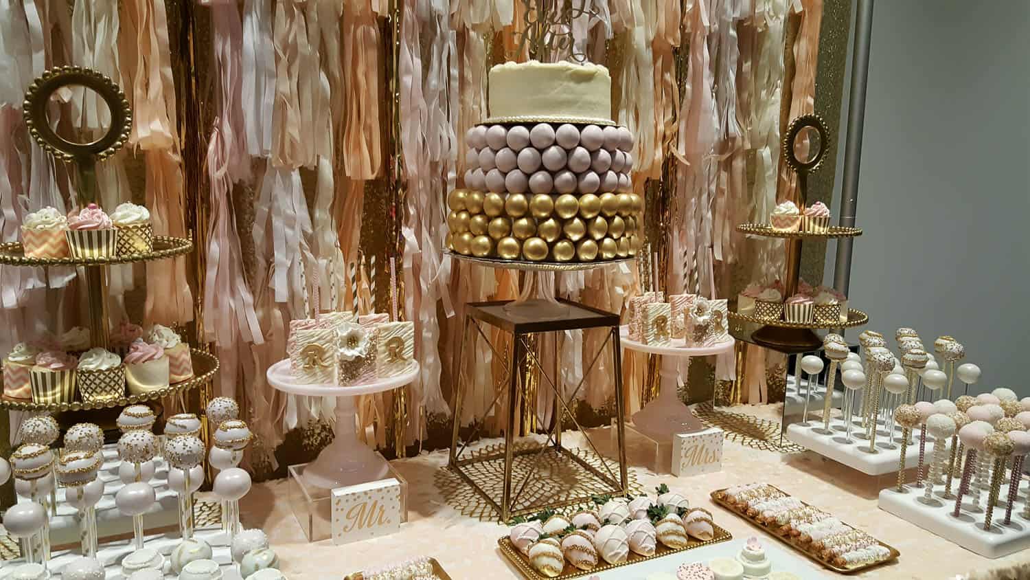 A dessert table with a white and gold tiered cake, cupcakes, cake pops, cookies, and pastries arranged on stands against a pink and gold ribbon backdrop.