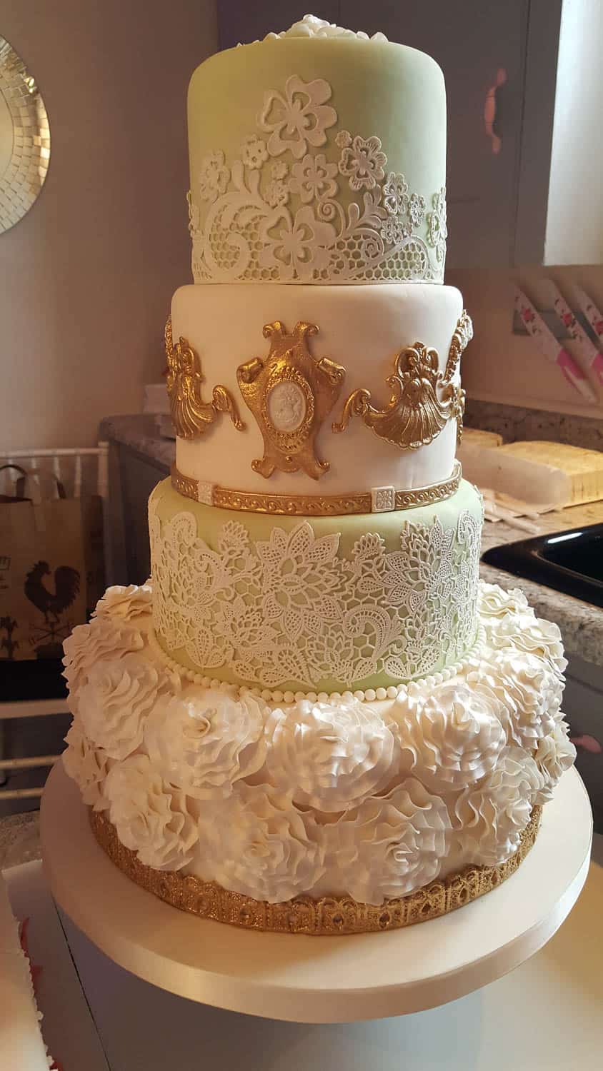 A four-tiered decorative cake with intricate lace designs, gold accents, and detailed white flower decorations on the bottom tier.