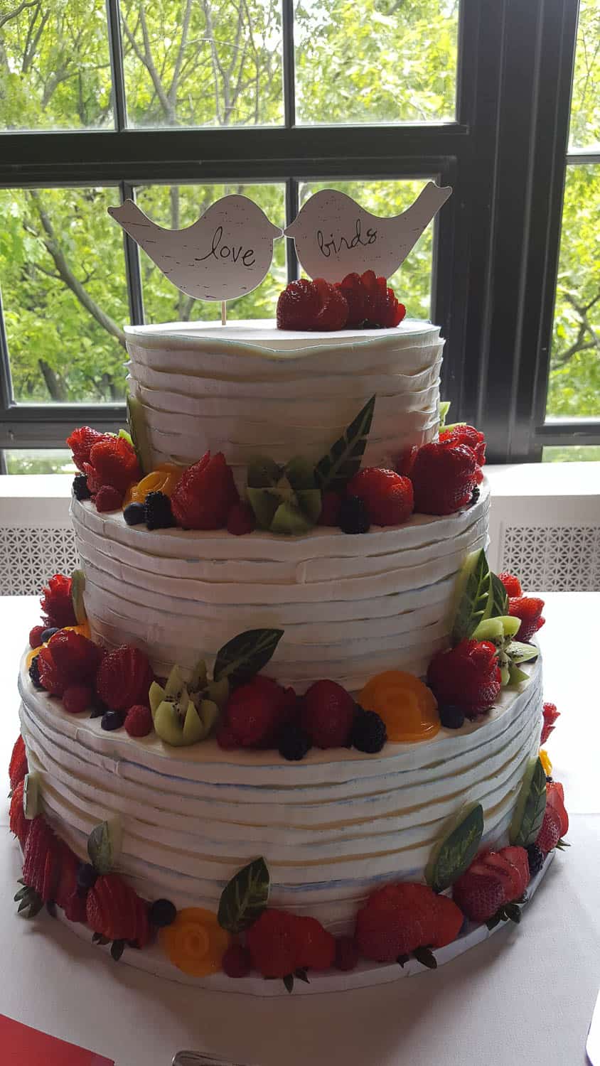 Three-tiered white frosted cake decorated with fresh fruit and topped with two paper birds labeled "love" and "birds," displayed in front of a window.