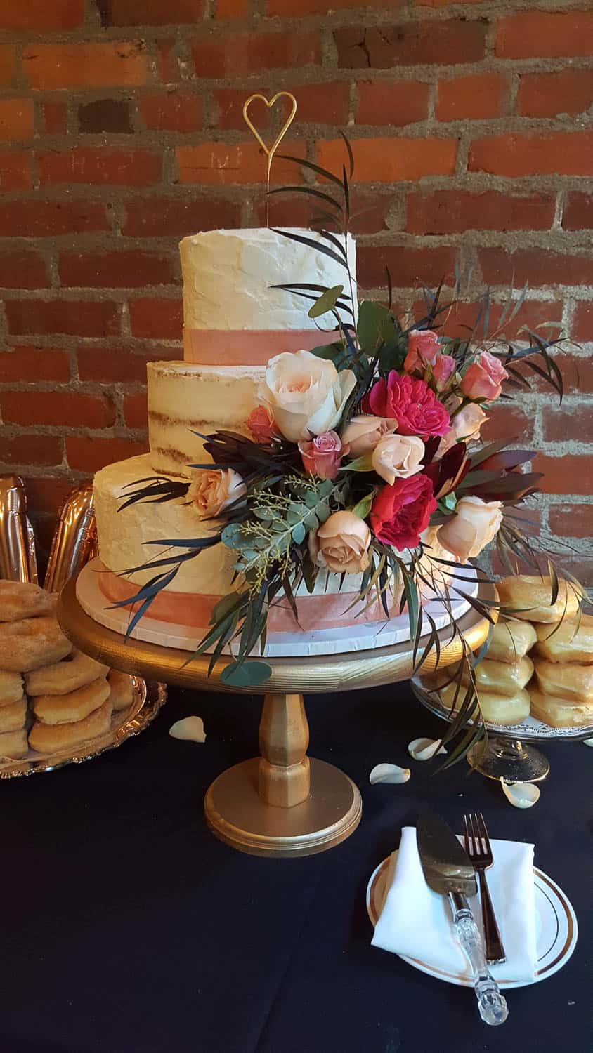 Three-tiered white cake with pink ribbon accents, decorated with fresh flowers and greenery, displayed on a wooden stand with donuts and tableware nearby.