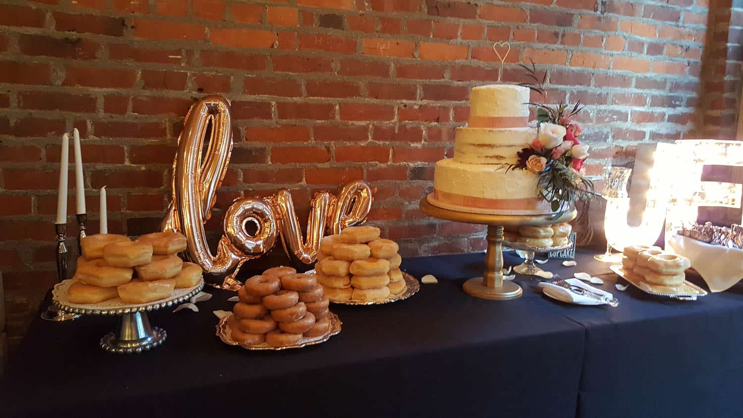 A dessert table with stacks of donuts, a tiered wedding cake with flowers, a "love" balloon, and candles, set against a brick wall.