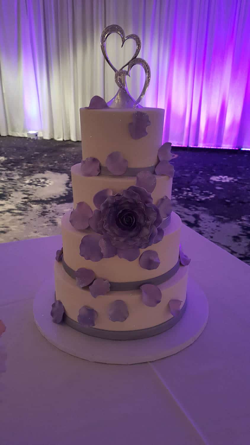 Four-tier white cake decorated with purple flower petals and a large purple flower, topped with a silver heart ornament, set against a purple-lit backdrop.