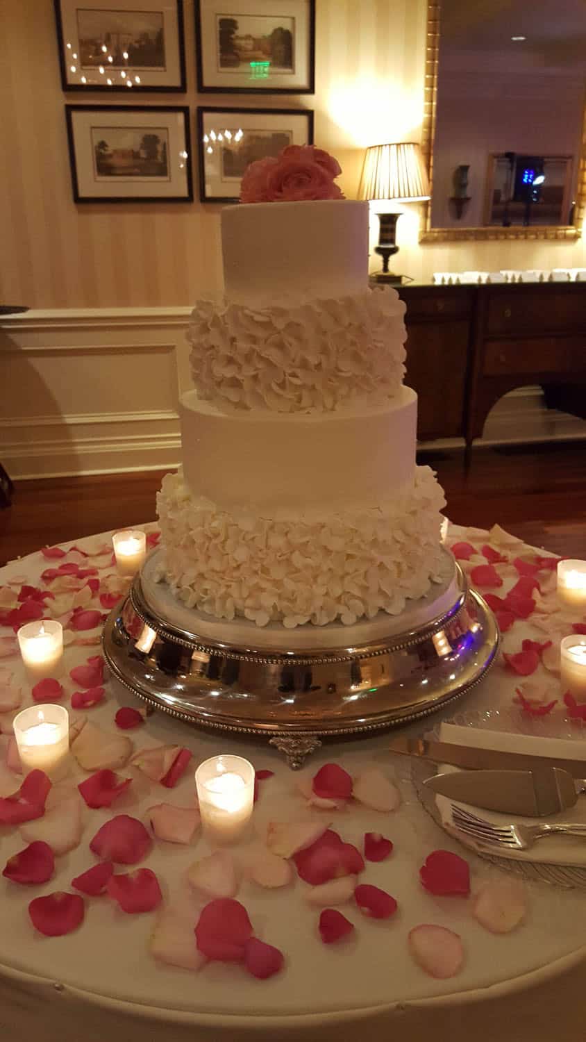 Four-tier white wedding cake with ruffled icing, topped with pink flowers, on a silver stand surrounded by rose petals and lit candles on a round table.