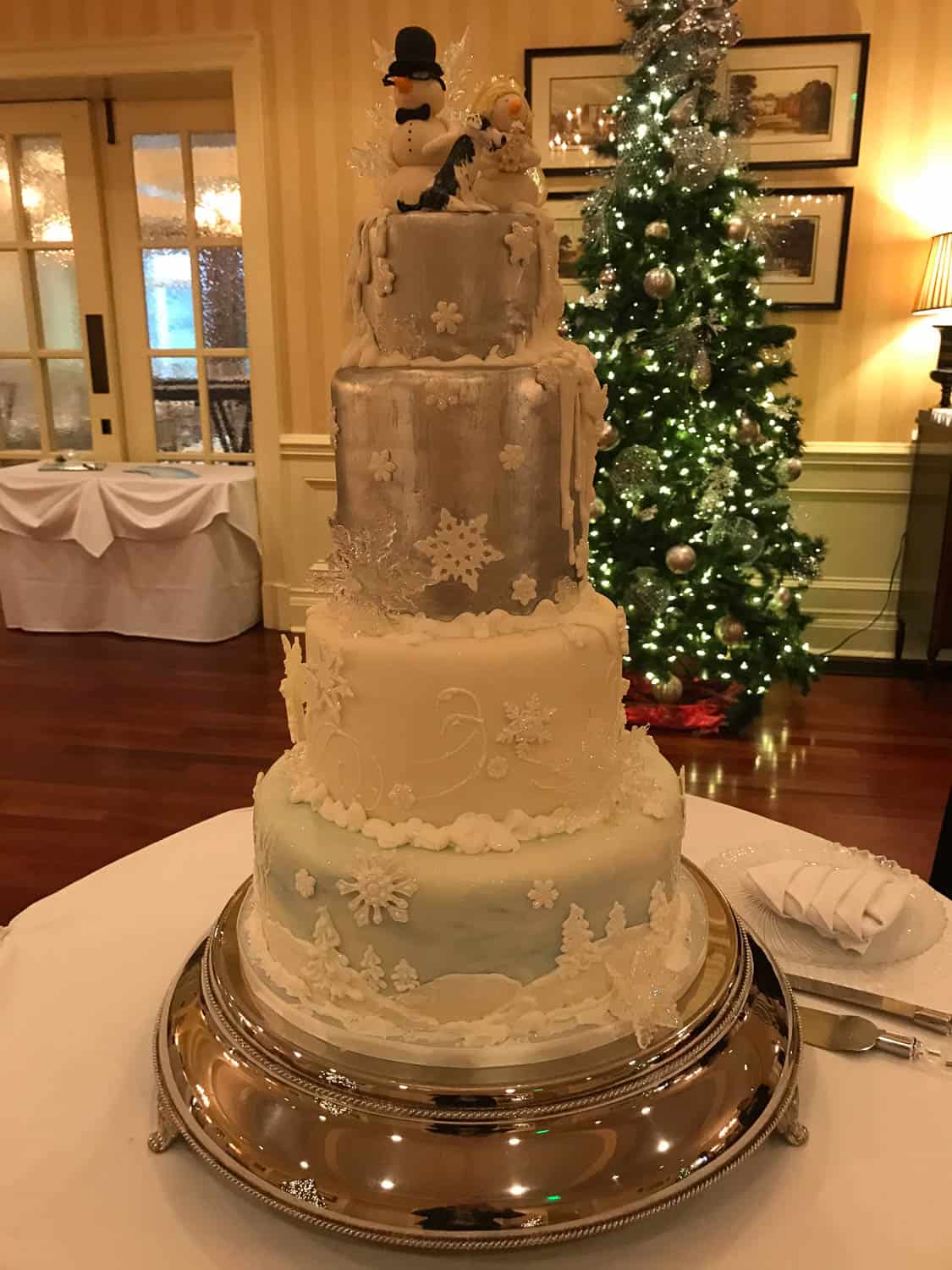 A four-tiered winter-themed cake with snowflake decorations and a snowman topper, displayed on a silver tray in front of a decorated Christmas tree.