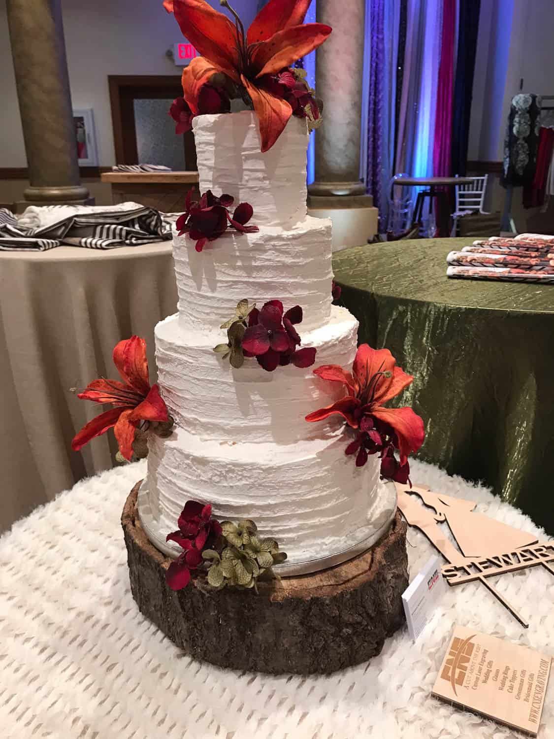 A four-tiered white cake decorated with red and orange flowers, displayed on a wooden slab base at an indoor event.