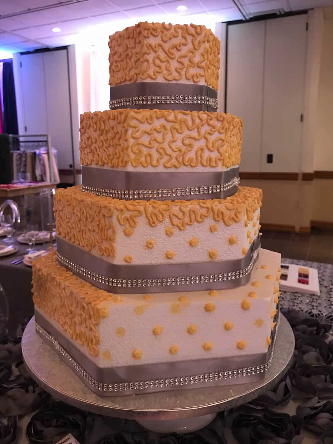 Four-tiered hexagonal cake with yellow piped icing, silver ribbon, and dot decorations, displayed indoors on a round silver base.