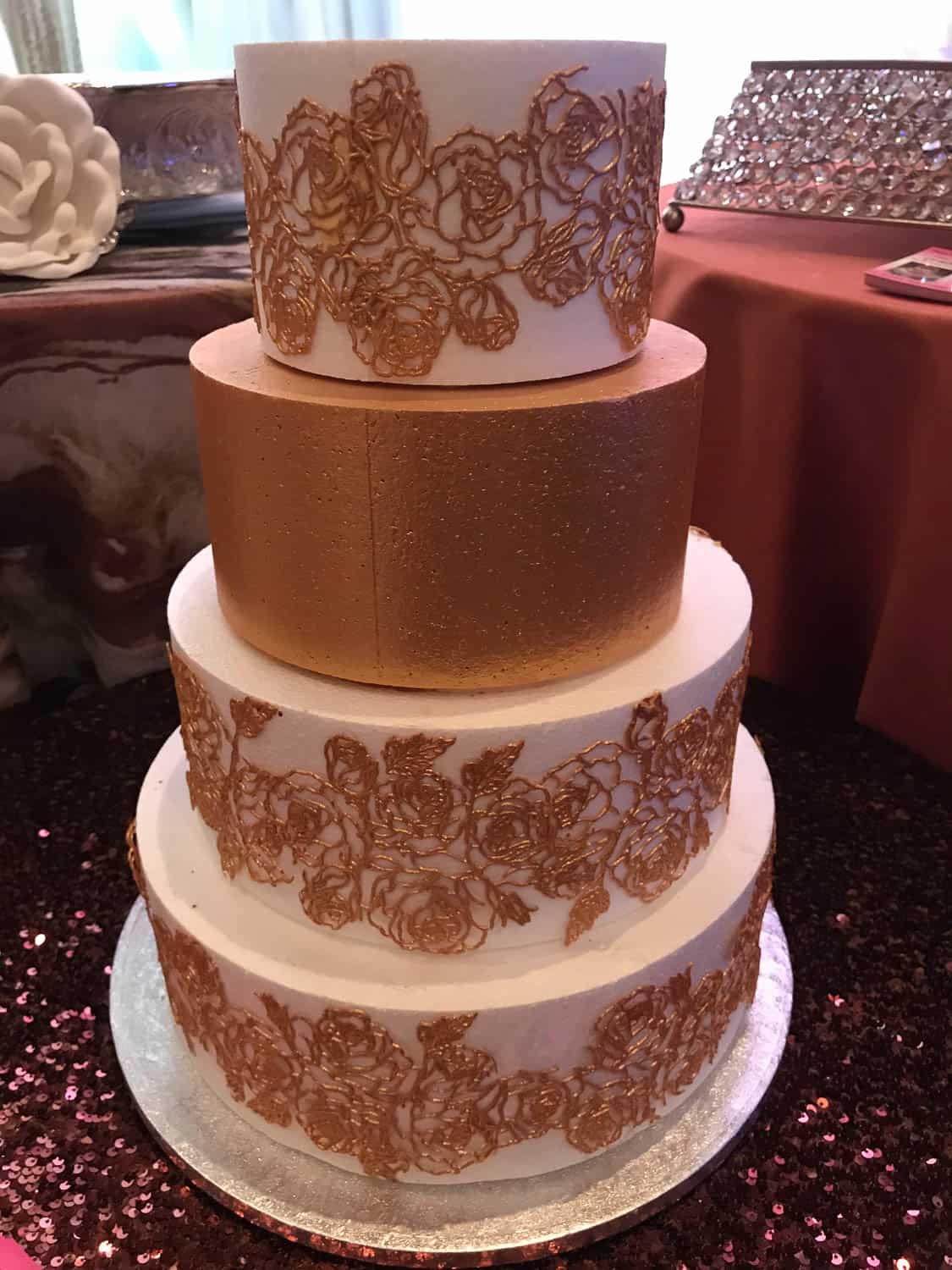 A four-tiered white cake with metallic gold middle tier and gold floral lace decorations on the top and bottom tiers, on a table with a sequin tablecloth.