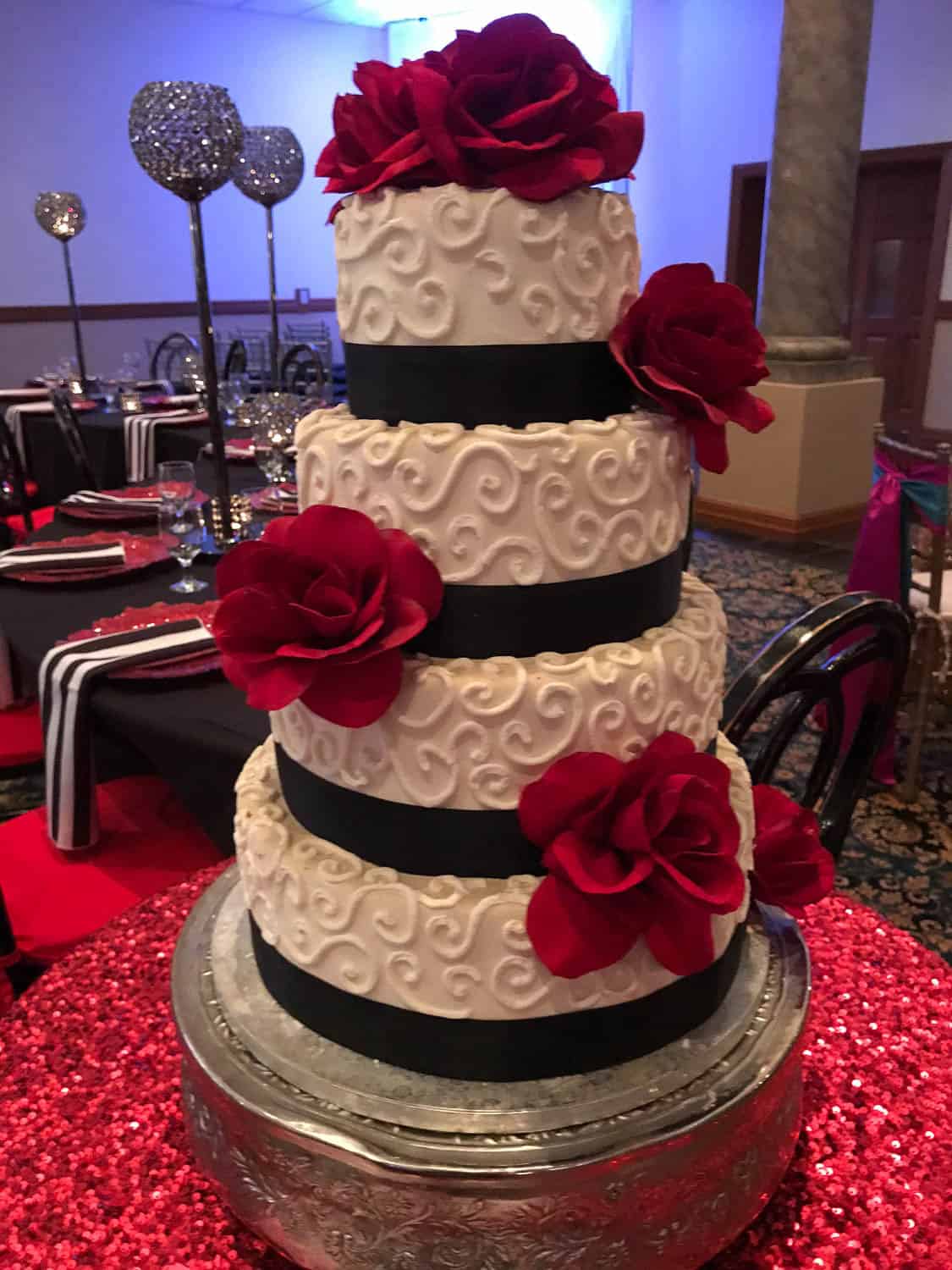 Four-tier white wedding cake with black ribbon bands, red fondant roses, and a swirl pattern, displayed on a silver stand at a decorated event table.