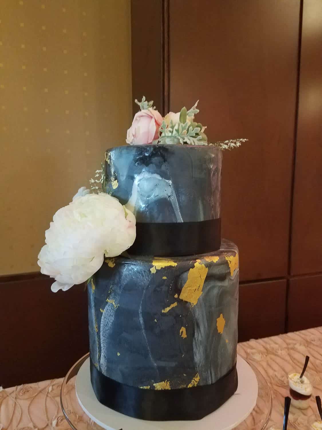Two-tiered cake with a dark marbled design, gold accents, black ribbon bands, and topped with white and pink flowers and greenery.