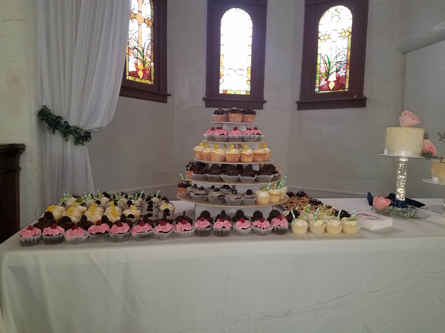 A tiered cupcake stand and a variety of decorated cupcakes are displayed on a white tablecloth in front of stained glass windows.