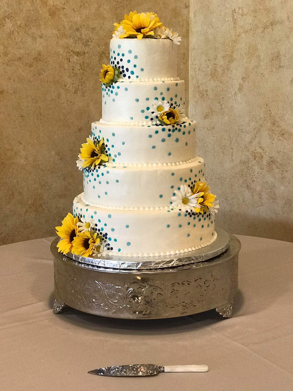 Five-tiered white cake decorated with blue polka dots and yellow artificial flowers, displayed on a silver stand with a cake knife in front.