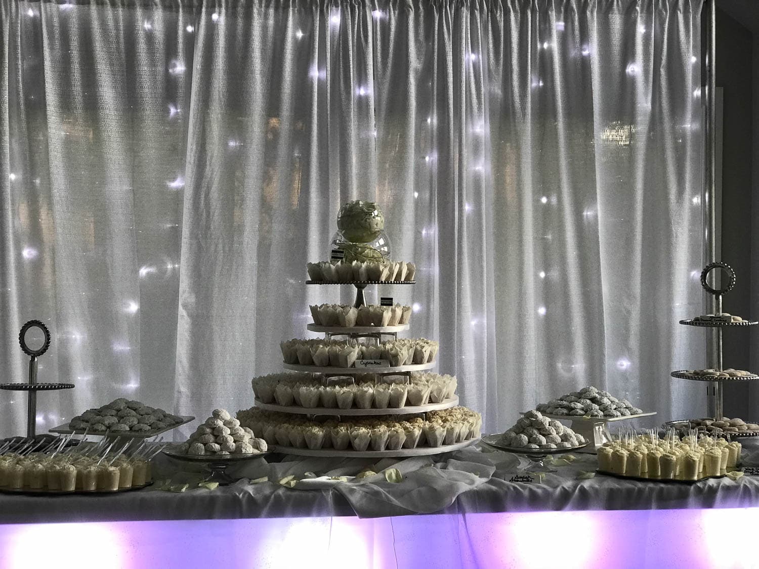 A tiered dessert stand with cupcakes, centered on a table, surrounded by assorted white and beige sweets, against a backdrop of sheer curtains and string lights.