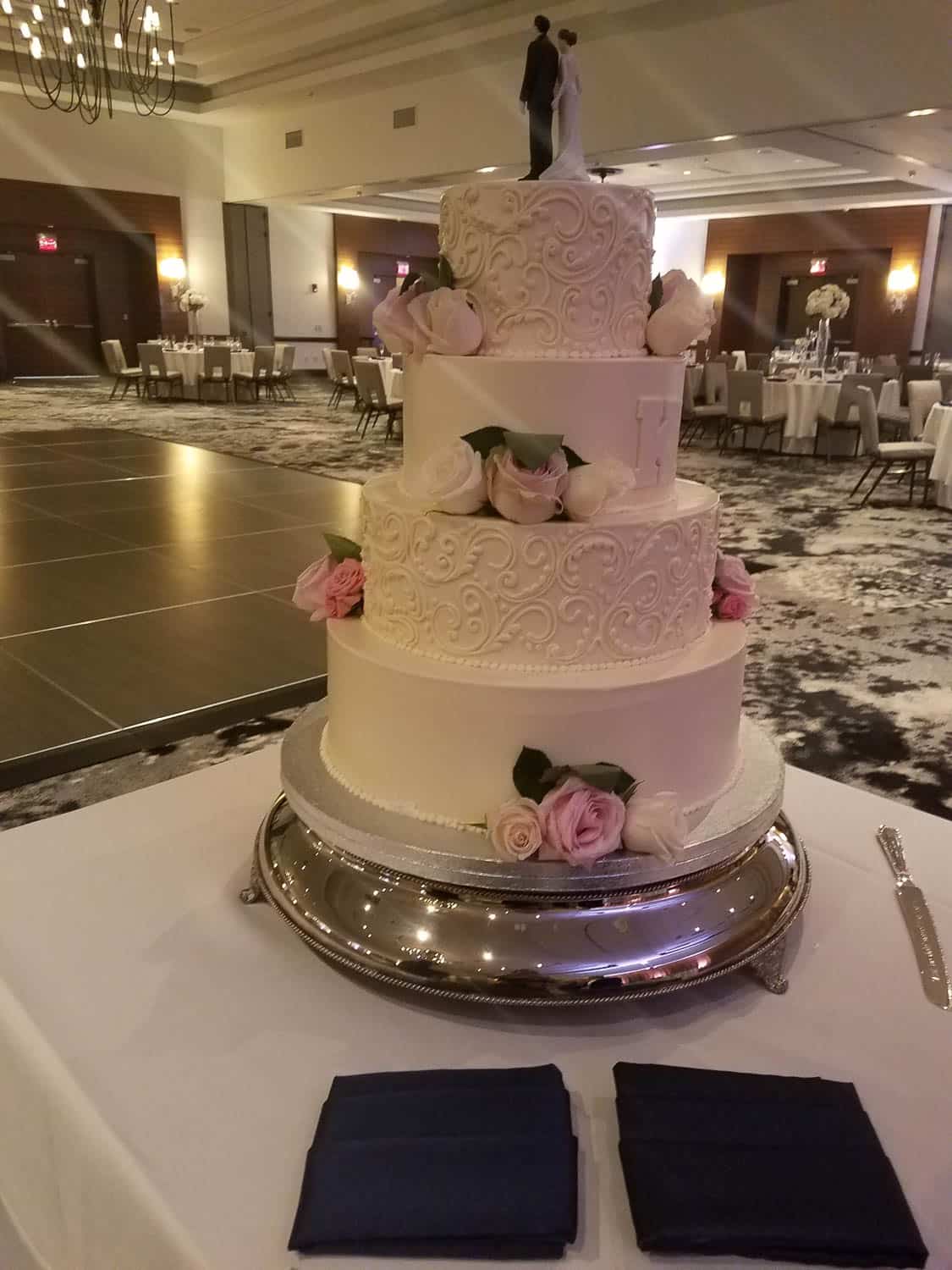 A three-tiered white wedding cake decorated with pink roses and a bride and groom topper sits on a silver tray in an elegant banquet hall.