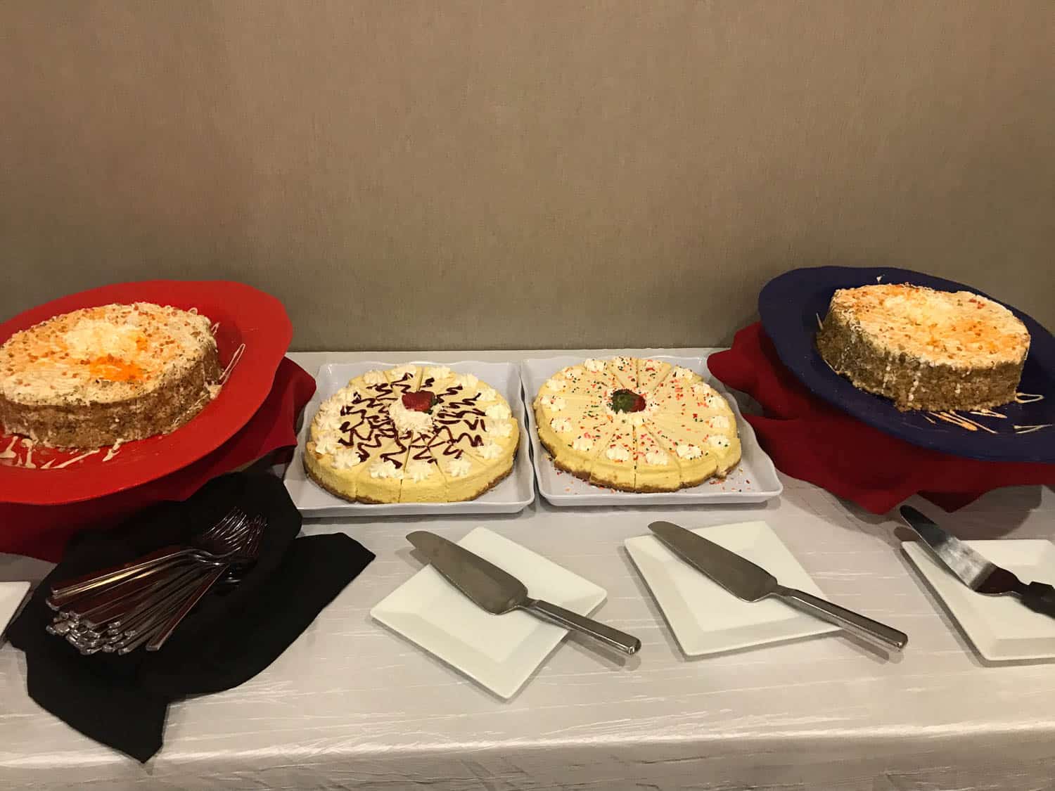 Four cheesecakes with various toppings are displayed on a table with serving utensils, plates, and forks arranged alongside them.