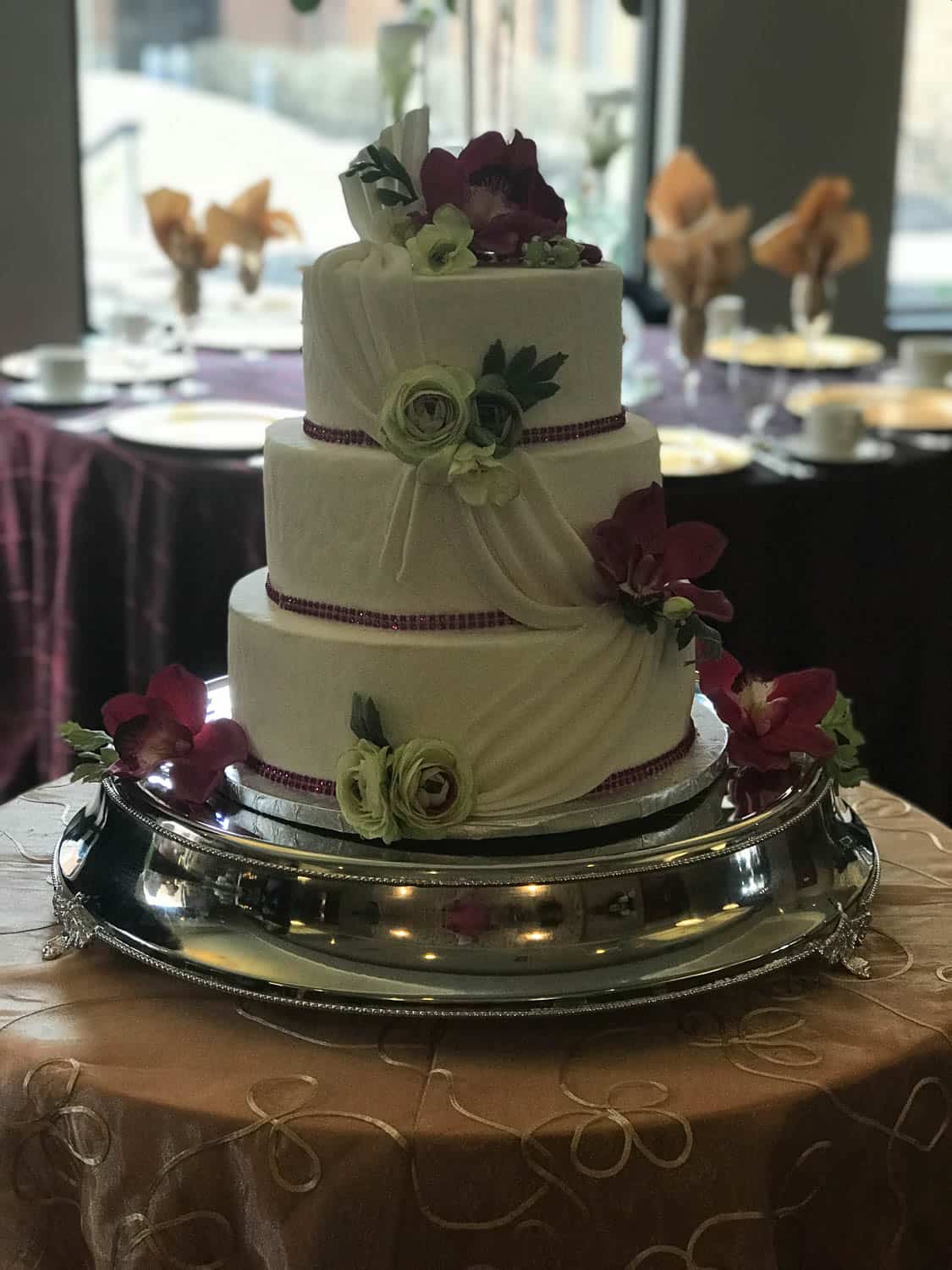 Three-tier white cake decorated with purple and green flowers, displayed on a silver stand atop a round table with a beige tablecloth.