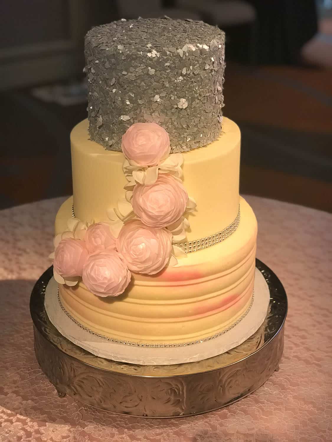 Three-tiered cake with gray textured top, smooth yellow tiers, pink and white fondant flowers, and silver bands, displayed on a round stand.