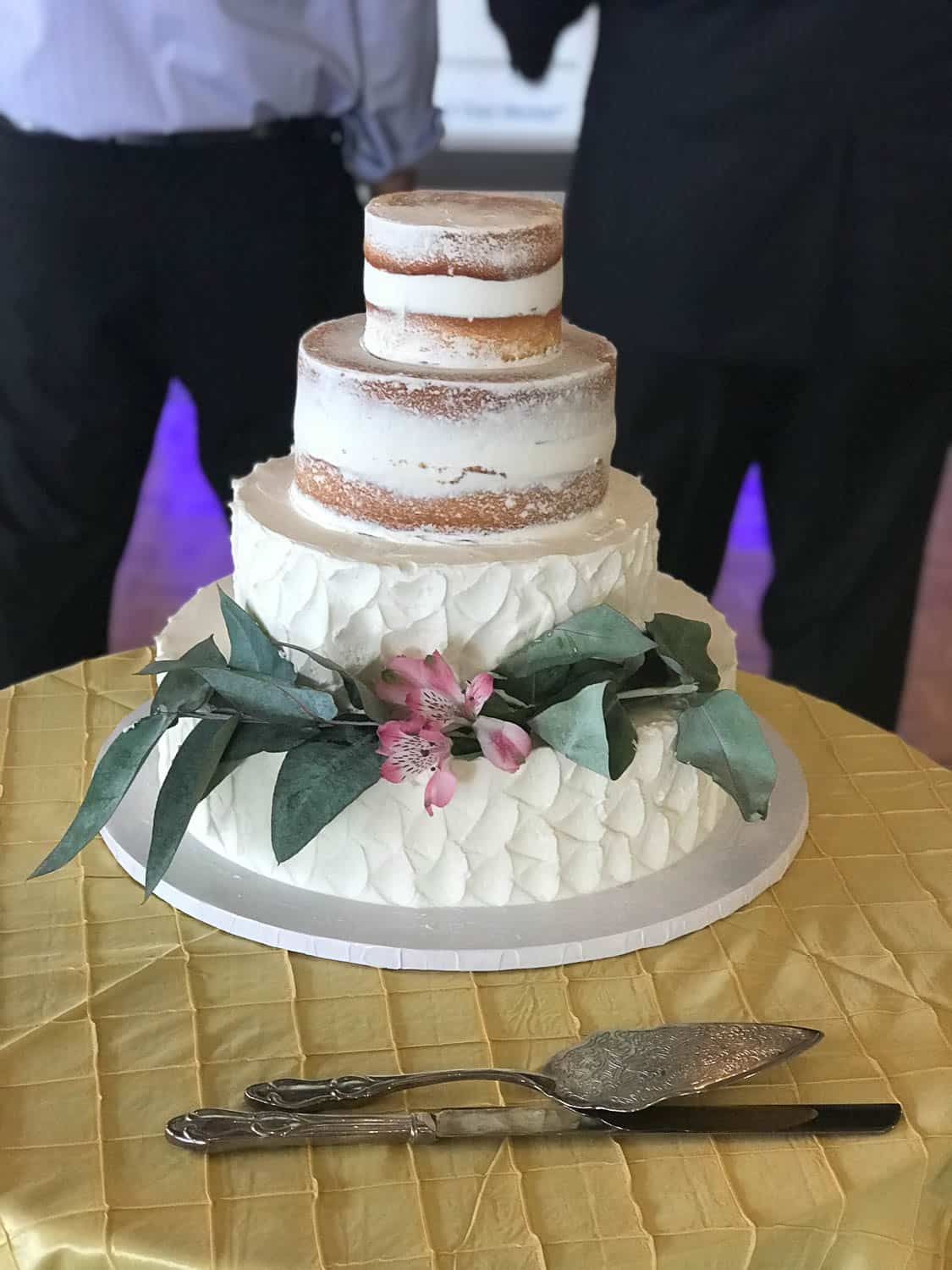 A three-tiered semi-naked cake with white icing, greenery, and pink flowers sits on a round table with a yellow cloth; two people stand in the background.
