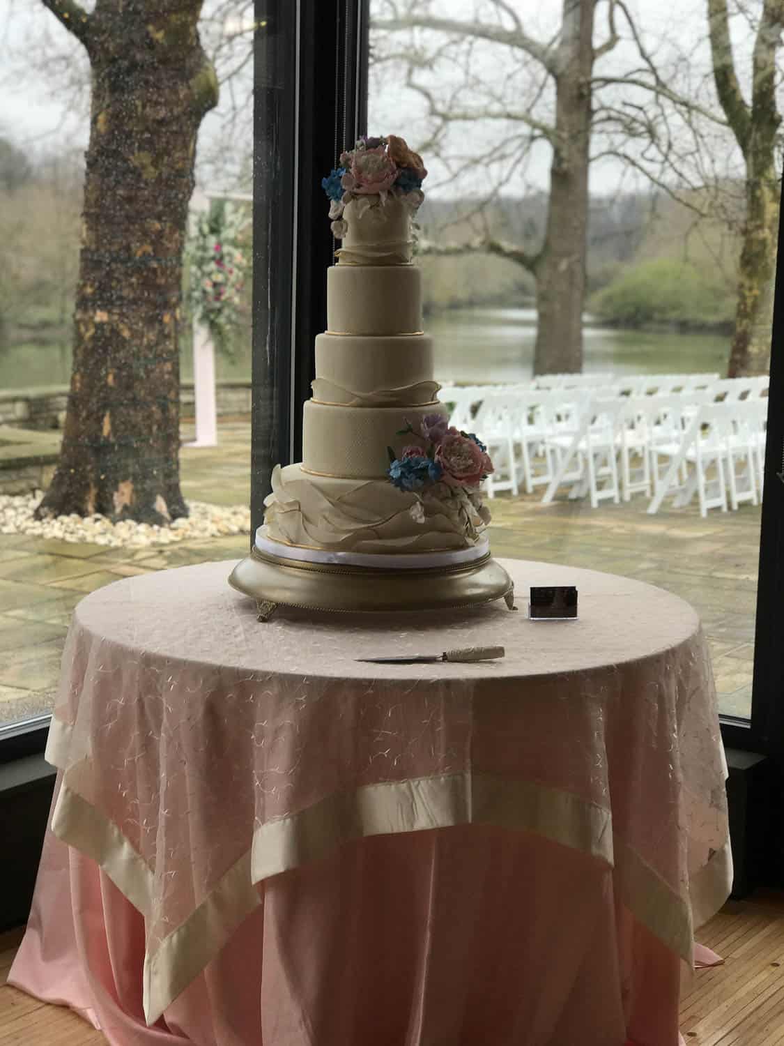 Tall white tiered wedding cake with floral decorations on a round table with pink and white cloth, near windows overlooking a patio with white chairs.