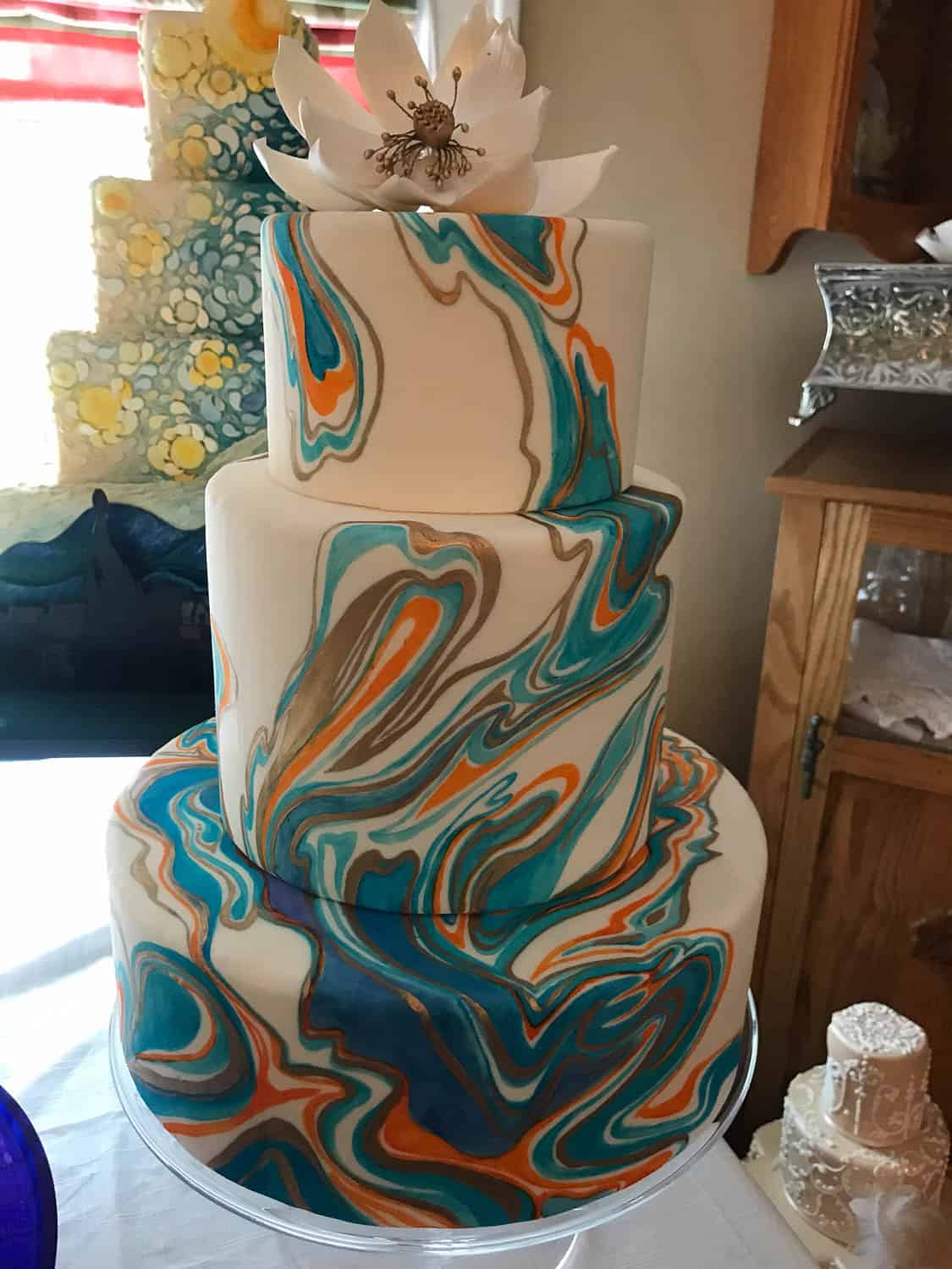Three-tiered cake with marbled fondant in blue, orange, brown, and white, topped with a white flower decoration.