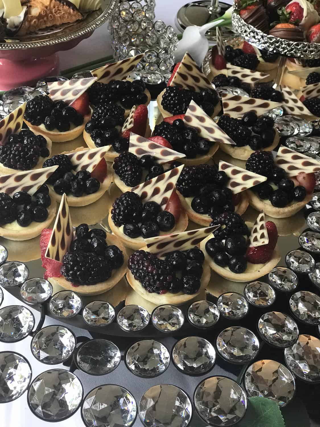 Mini fruit tarts topped with blackberries, blueberries, strawberries, and white chocolate pieces arranged on a tray decorated with faux crystals.