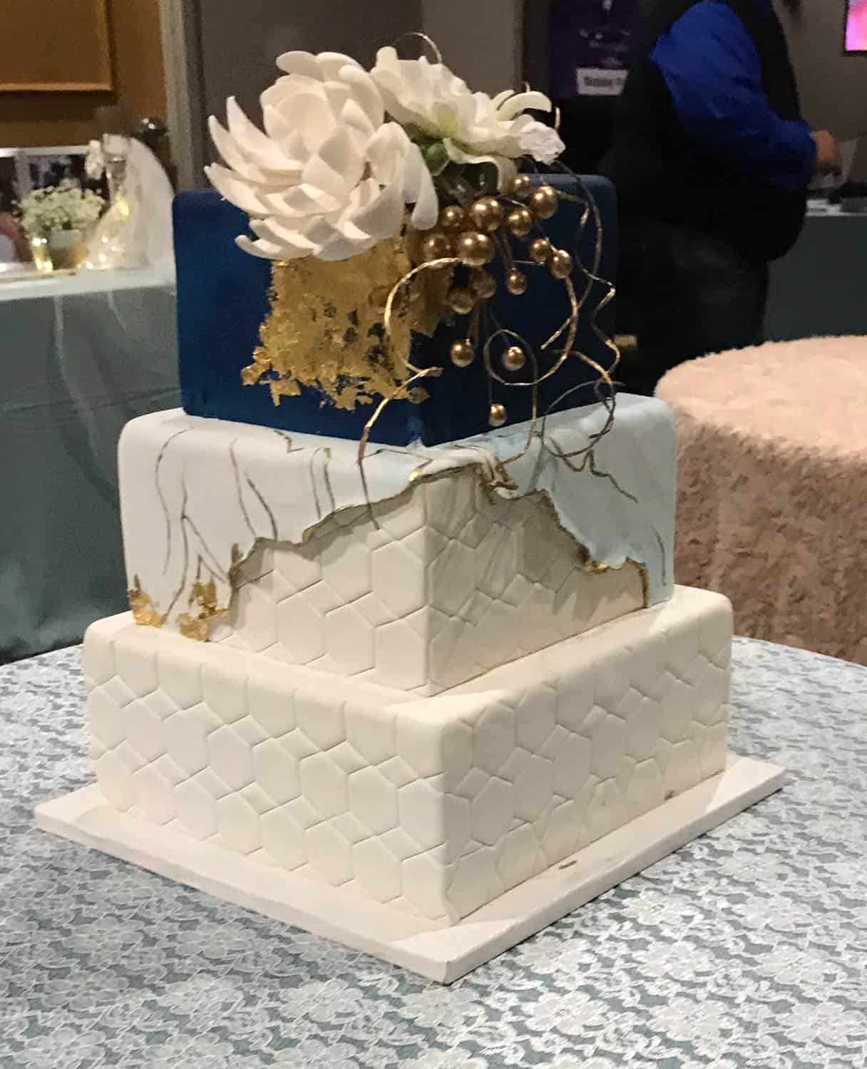 Three-tier square cake with geometric patterns, gold accents, and a large white flower on top, set on a lace-covered table.