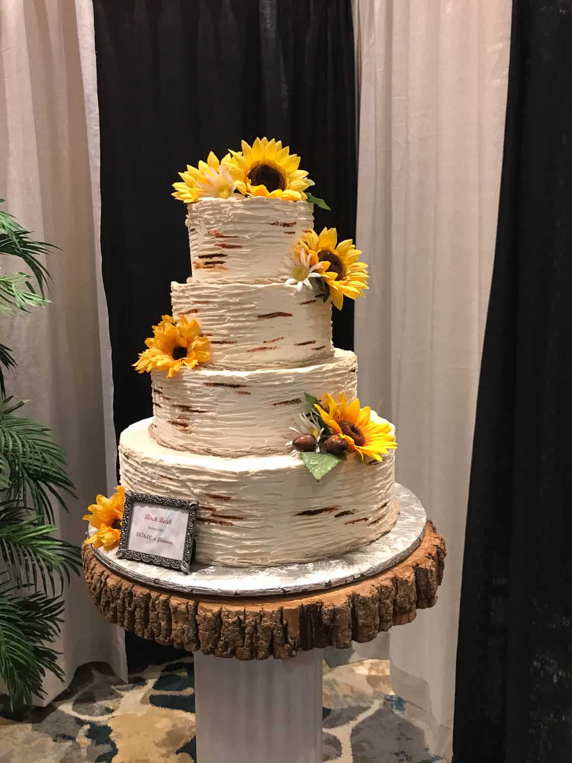 Three-tiered white cake with rustic, birch-like texture, decorated with sunflowers, displayed on a wood slice stand.