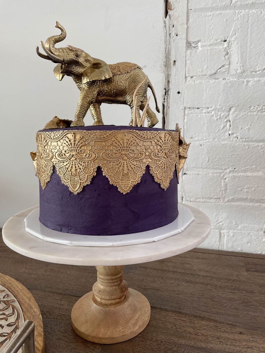 A round cake with purple frosting and gold lace decorations, topped with a gold elephant figurine, displayed on a wooden cake stand.