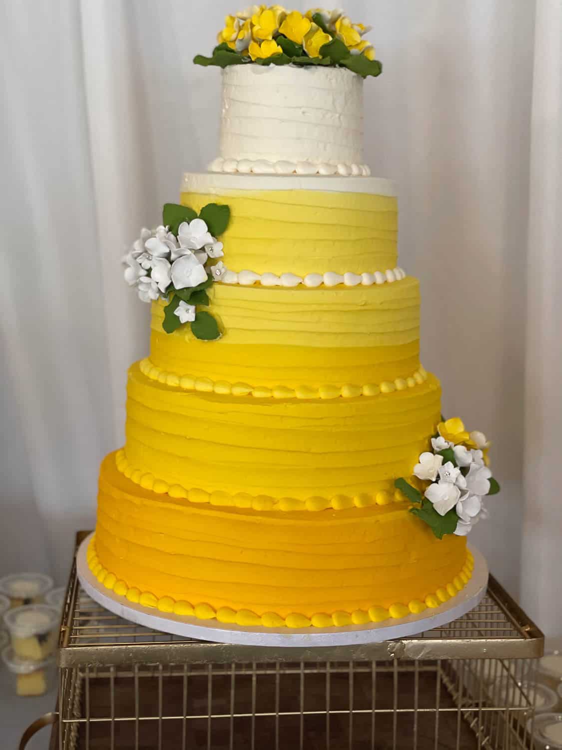 A five-tiered yellow and white cake decorated with sugar flowers, featuring gradient frosting from yellow at the base to white at the top.